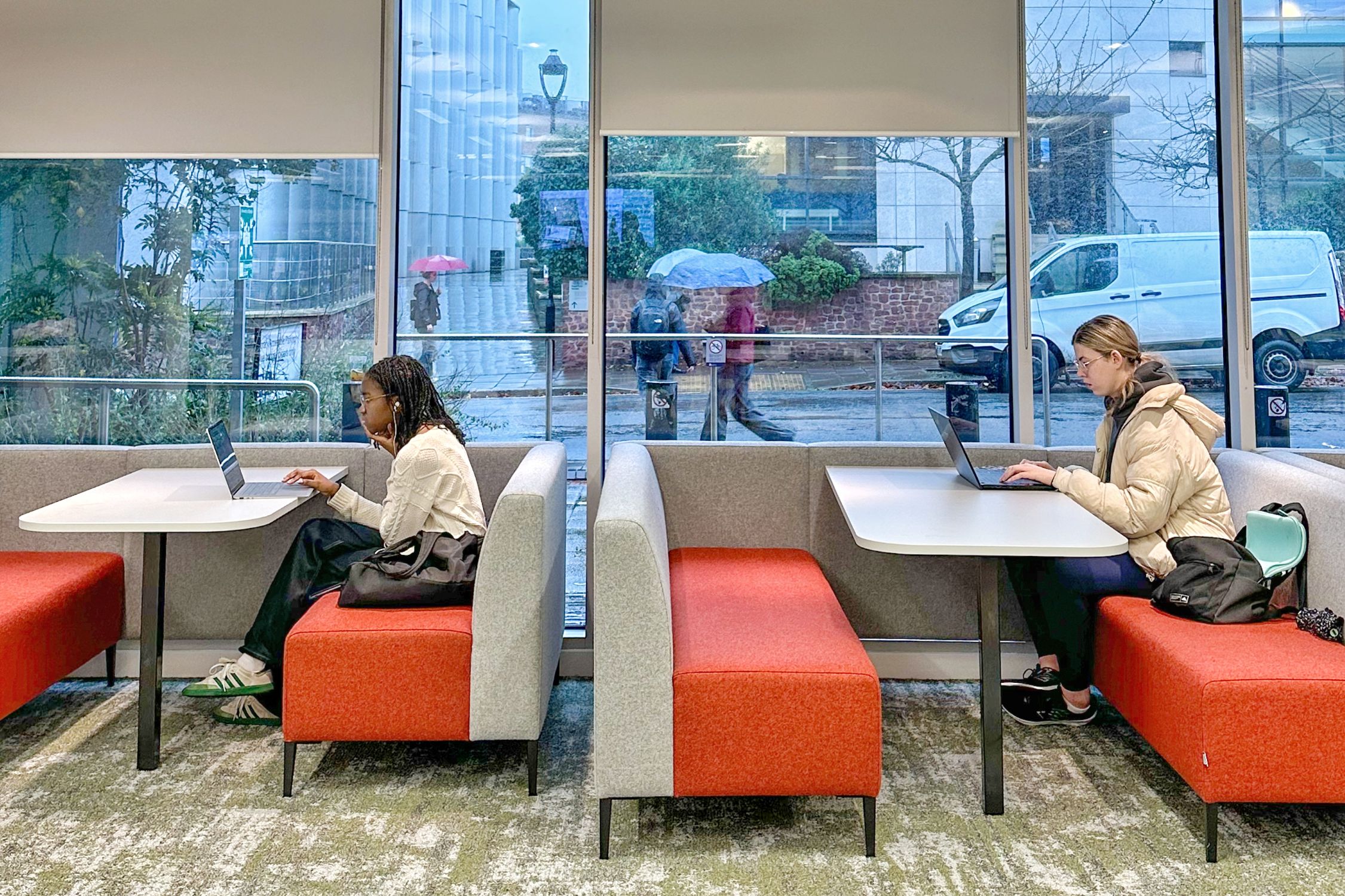 Two students sit on comfortable seating using laptops at tables in front of large windows